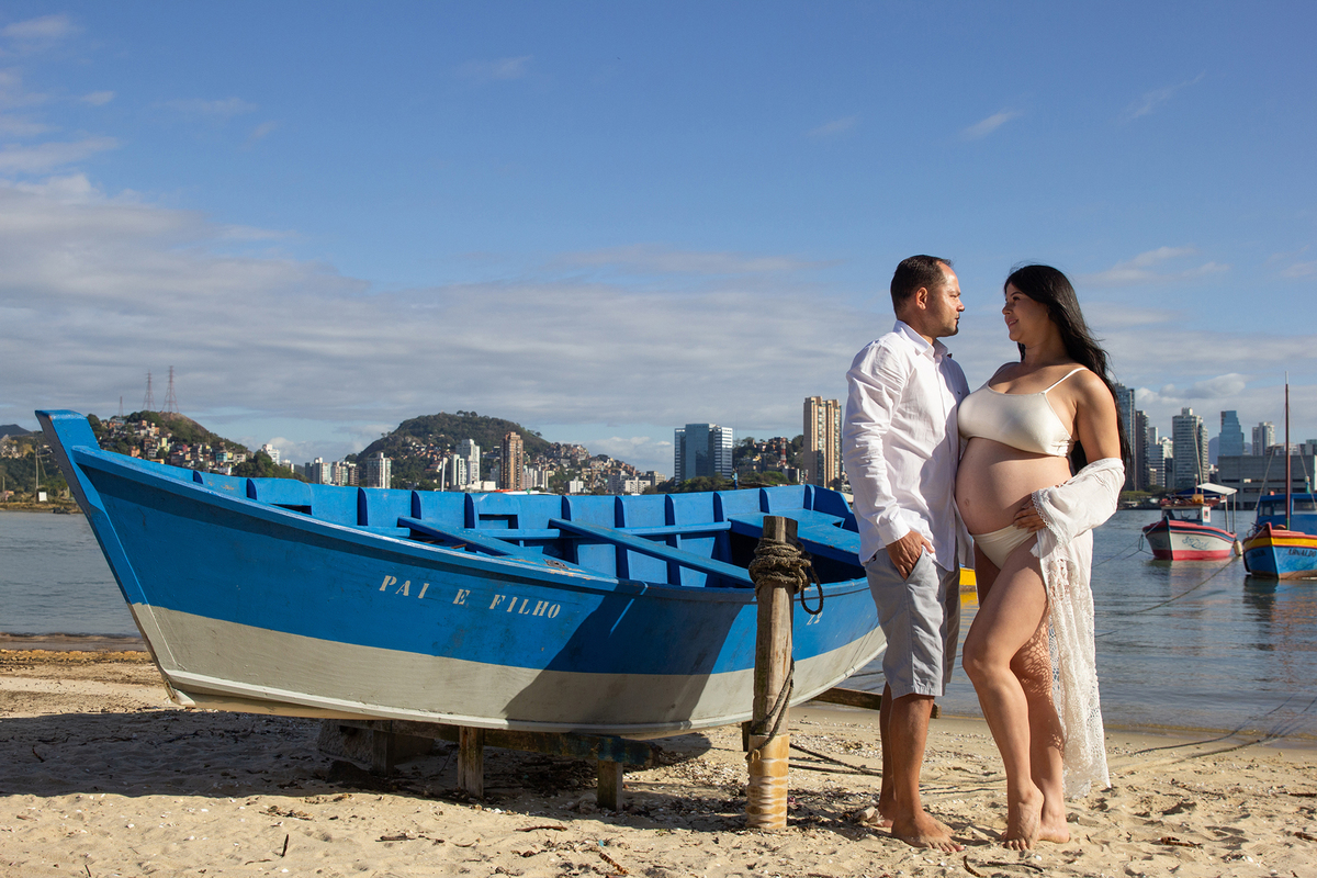Gestante posa para foto com marido, com as mãos na barriga. na areia, ao lado do barquinho, ao fundo o mar, barcos 