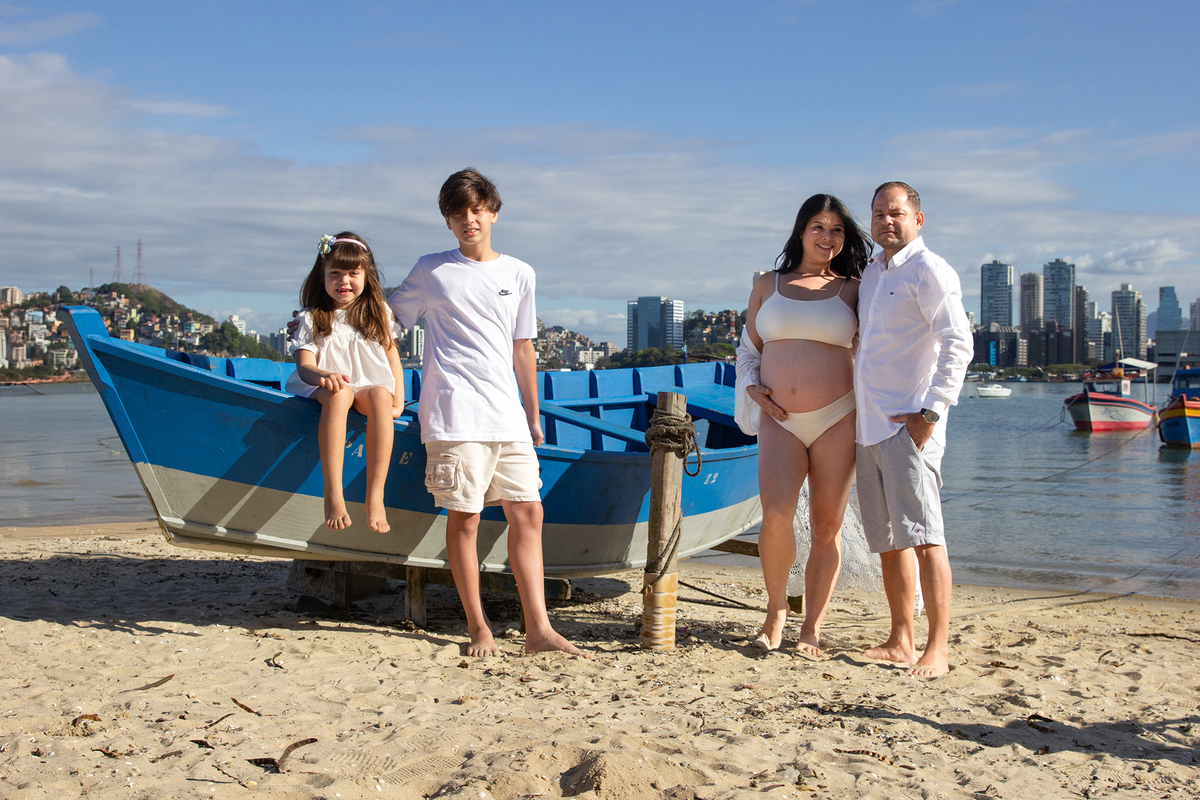 Gestante e família posa para foto, com as mãos na barriga. na areia, ao lado do barquinho, ao fundo o mar, barcos 