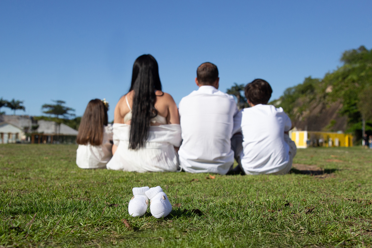 família sentada lado a lado, de costas, com destaque para sapatinha da bebê que vai chegar 