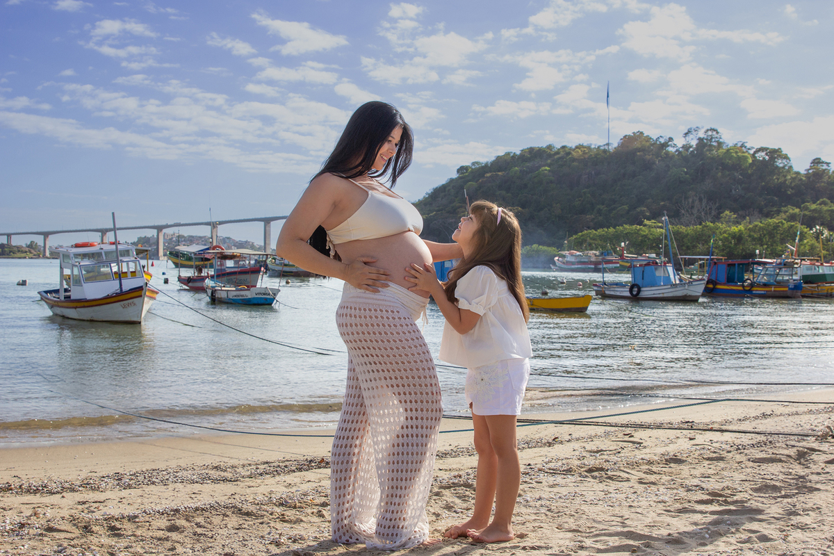 mãe e filha, se olhando e sorrindo, olhando para barriga, na praia, com mar e terceira ponte ao fundo 