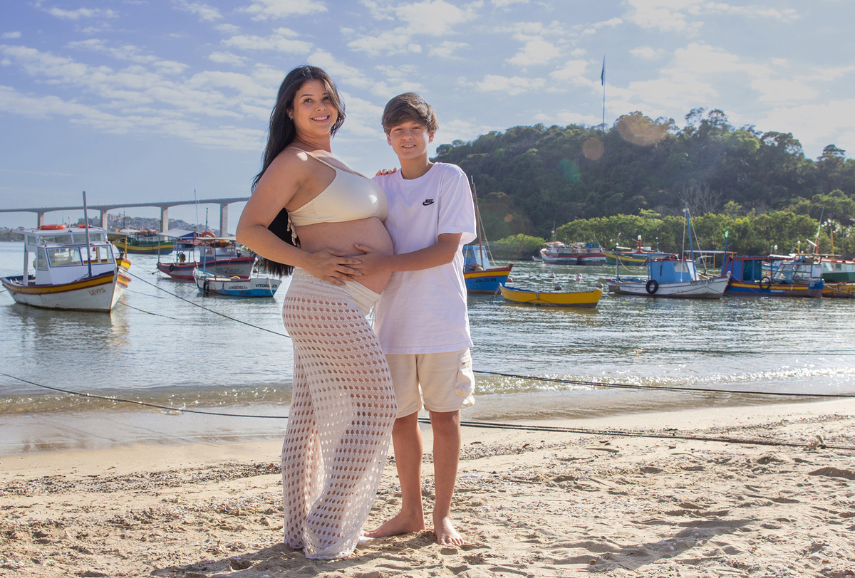 mãe e filho, olhando para barriga, na praia, com mar e terceira ponte ao fundo 