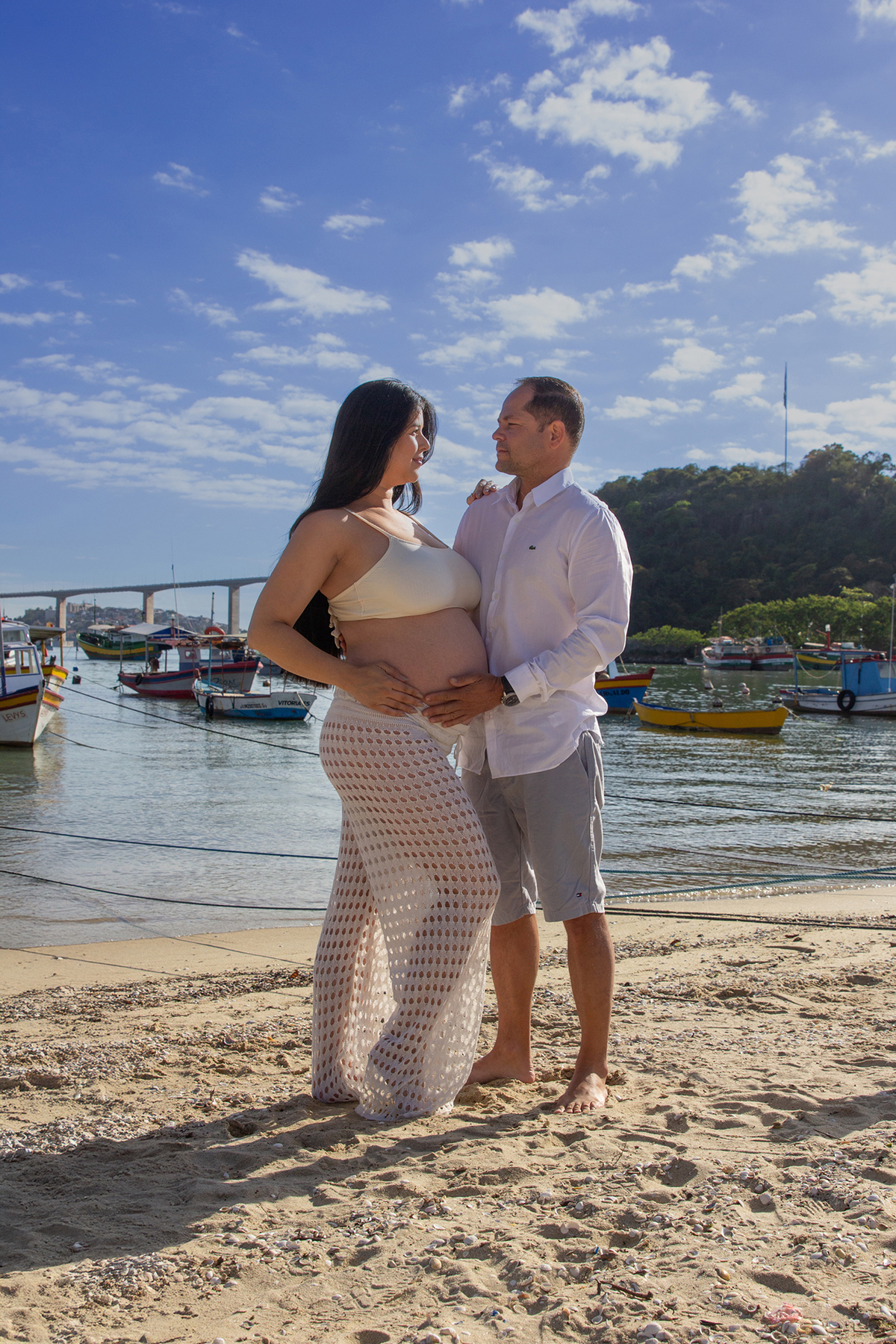 casal, olhando para barriga, na praia, com mar e terceira ponte ao fundo 