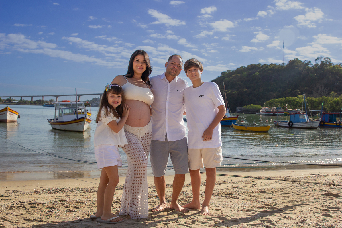 família abraçada posando, na praia, com mar e terceira ponte ao fundo 