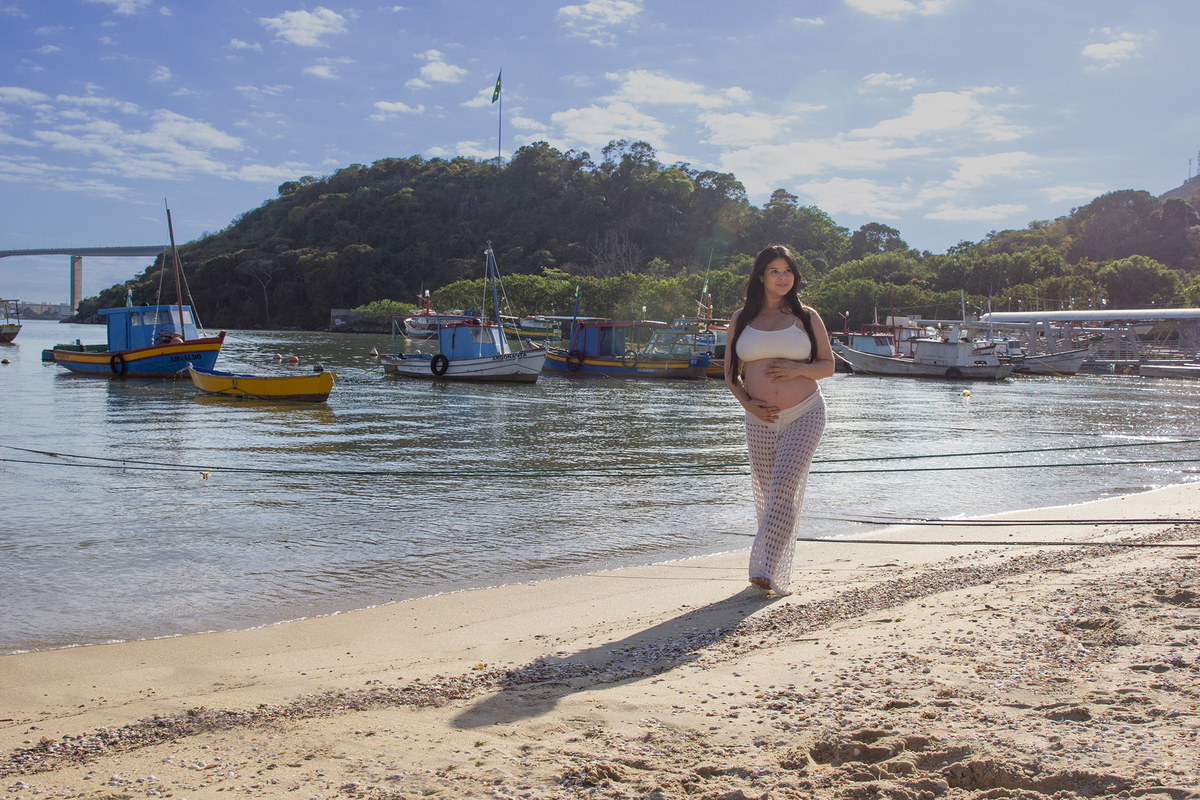 gestante caminha pela praia, com as mãos na barriga. ao fundo o mar, barcos e terceira ponte 