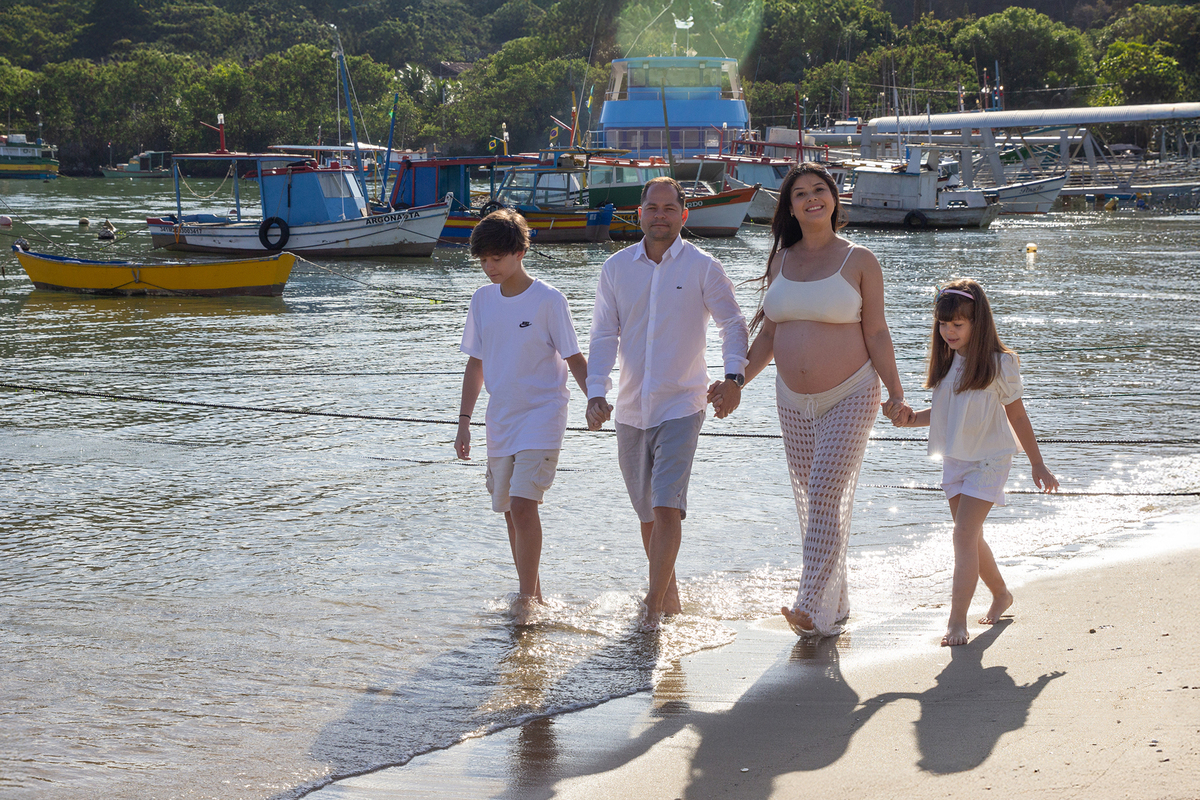 família caminha pela praia, com as mãos dadas. ao fundo o mar e barcos.