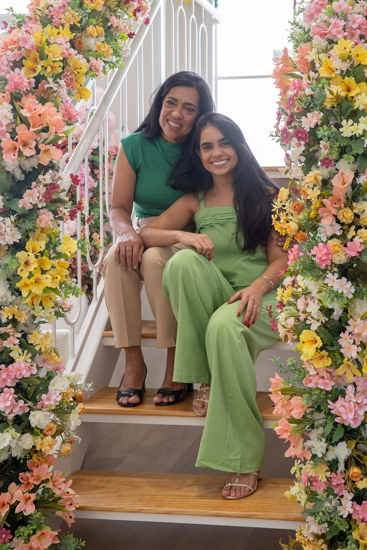 Mae e filha interagindo e sorrindo para foto juntas no cenário flores de dia das mães, sentadas na escada
