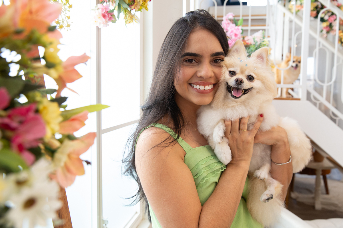 Spitz no colo da mãe de pet, ambas sorrindo, no cenário de dia das mães Flores. Outro spitz observa ao fundo 