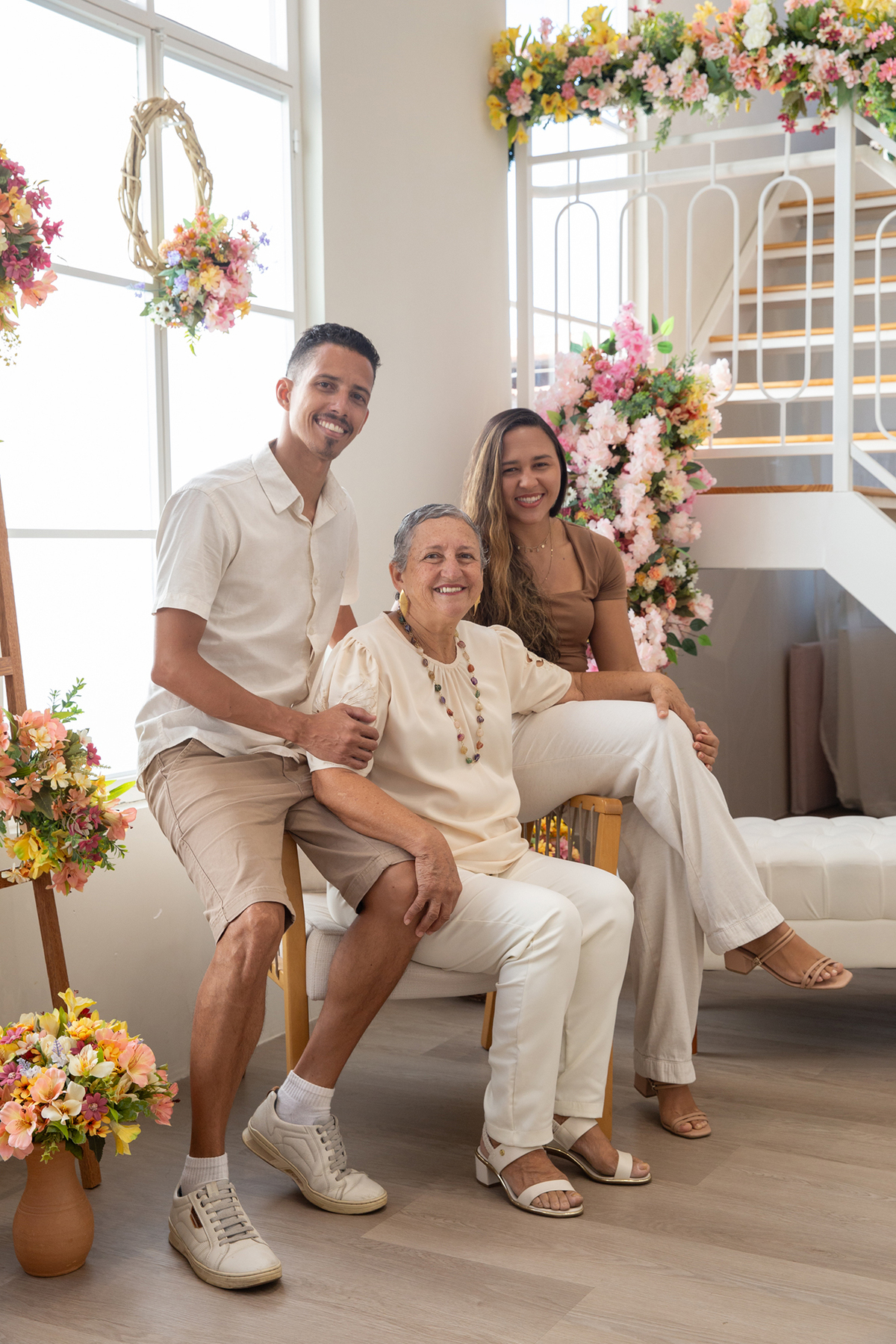 Mãe, filho e sua esposa, sorriem para câmera, no cenário flores do dia das mães