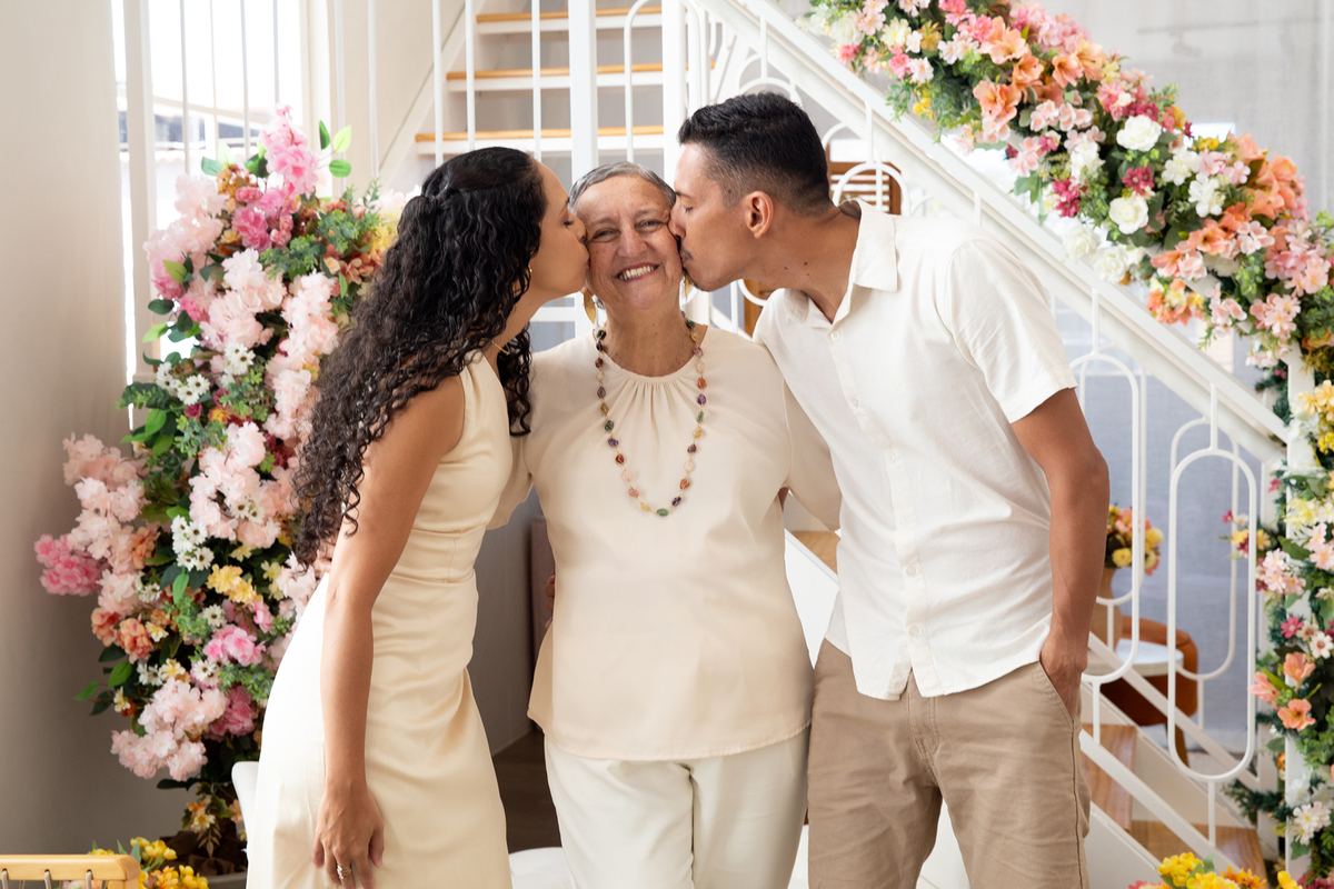 Mãe posa para foto recebendo beijo dos dois filhos, no cenário Flores de Dia das Mães