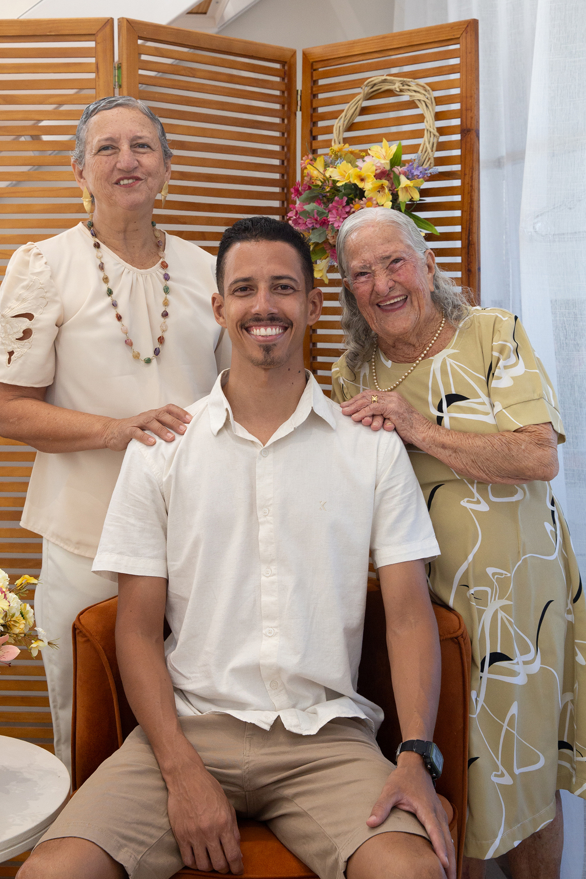 Mãe, filho e avó, sorriem para câmera, no cenário flores do dia das mães