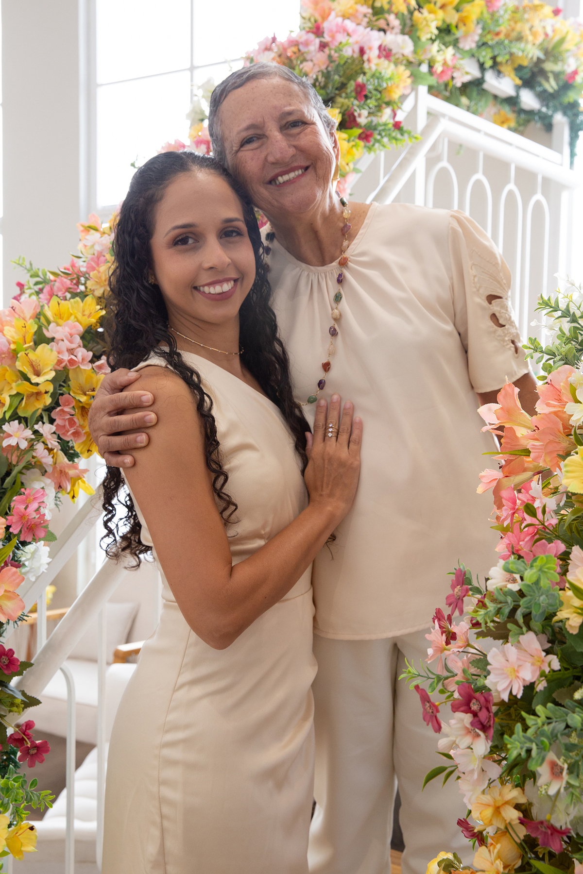 Mãe e filha, sorriem para câmera, no cenário flores do dia das mães