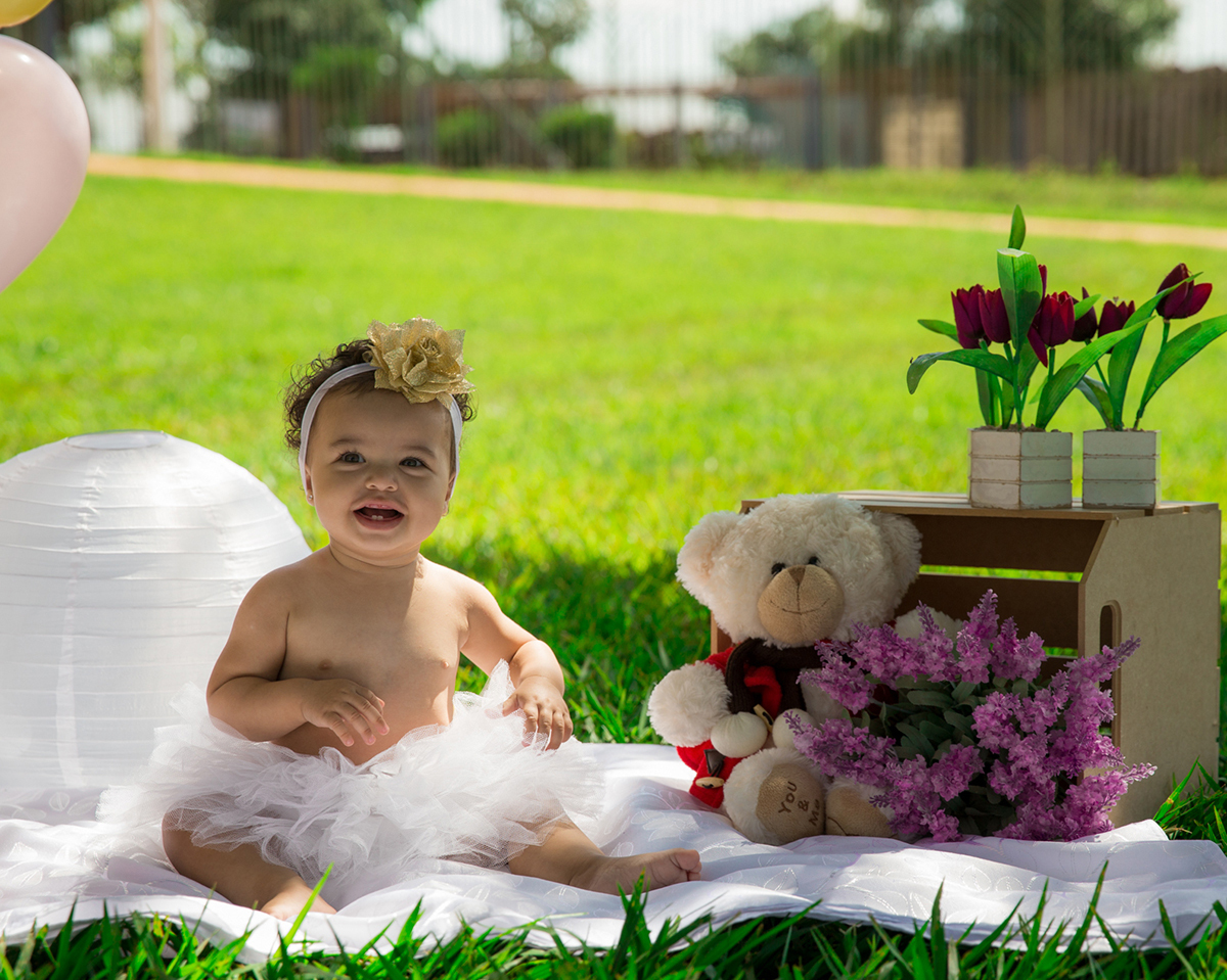 Bebê sentada no cenário, com balões e flores, vestida com saia de tutu, brinca e bate palmas, detalhes mais fechados