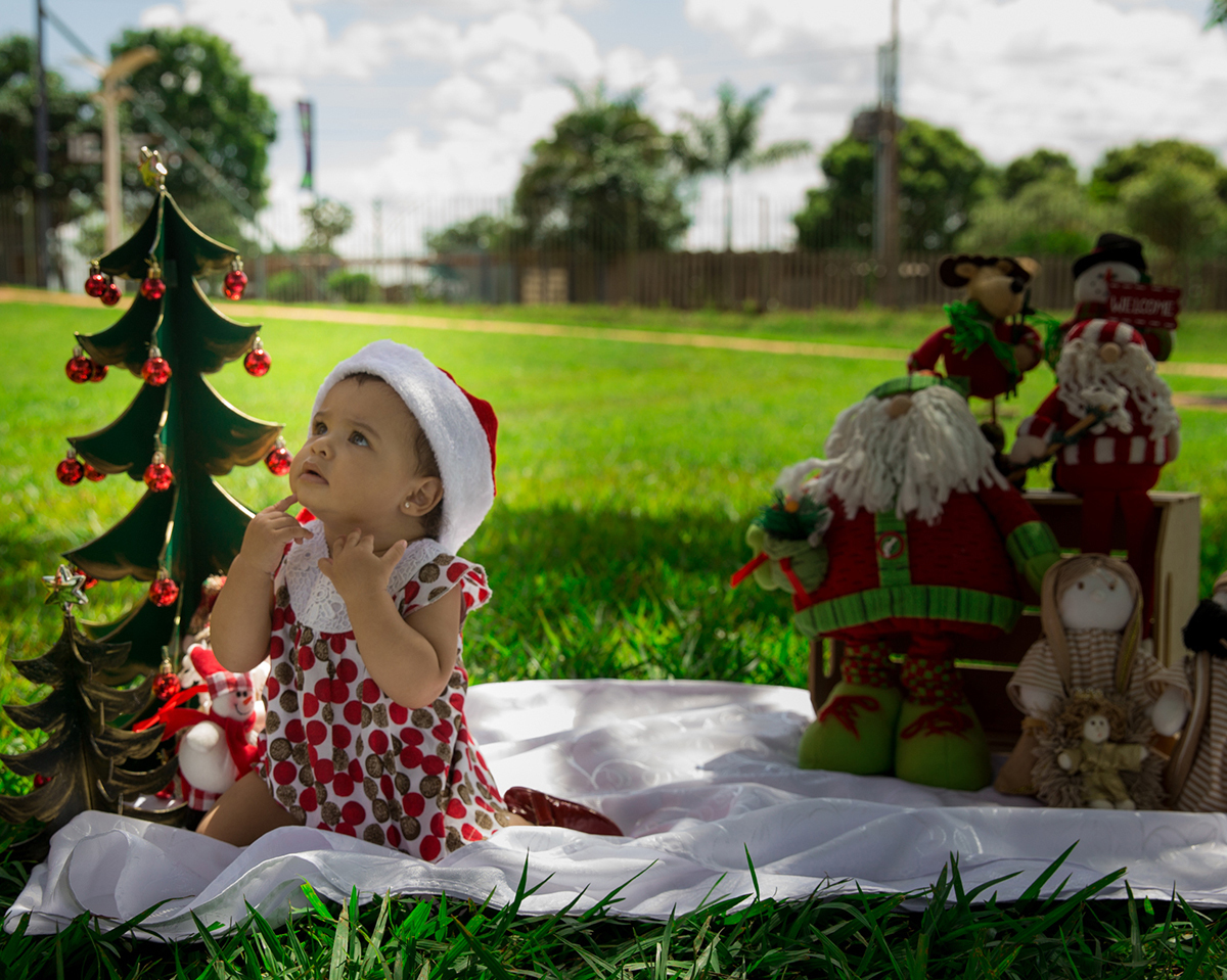 Bebê interage com cenário de natal, montado no gramado do parque, usa macacão vermelho e verde e touca de papai noel