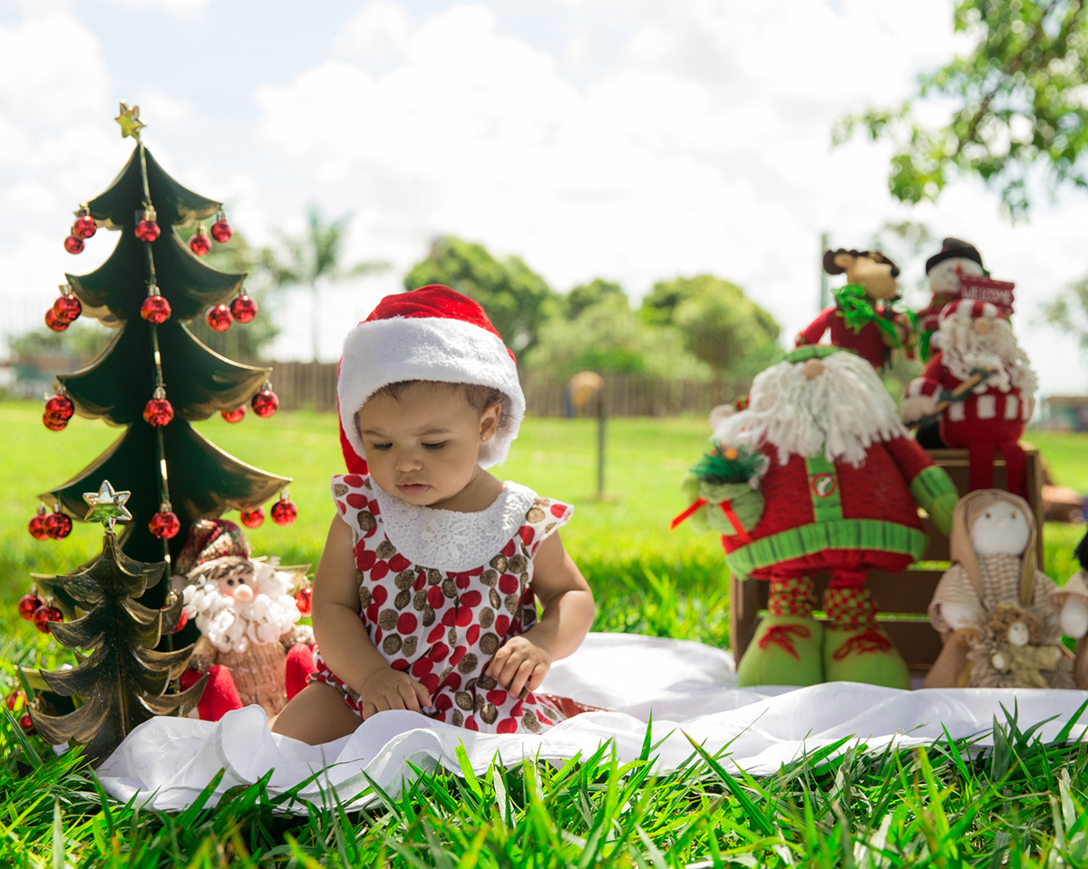 Bebê interage com cenário de natal, montado no gramado do parque, usa macacão vermelho e verde e touca de papai noel