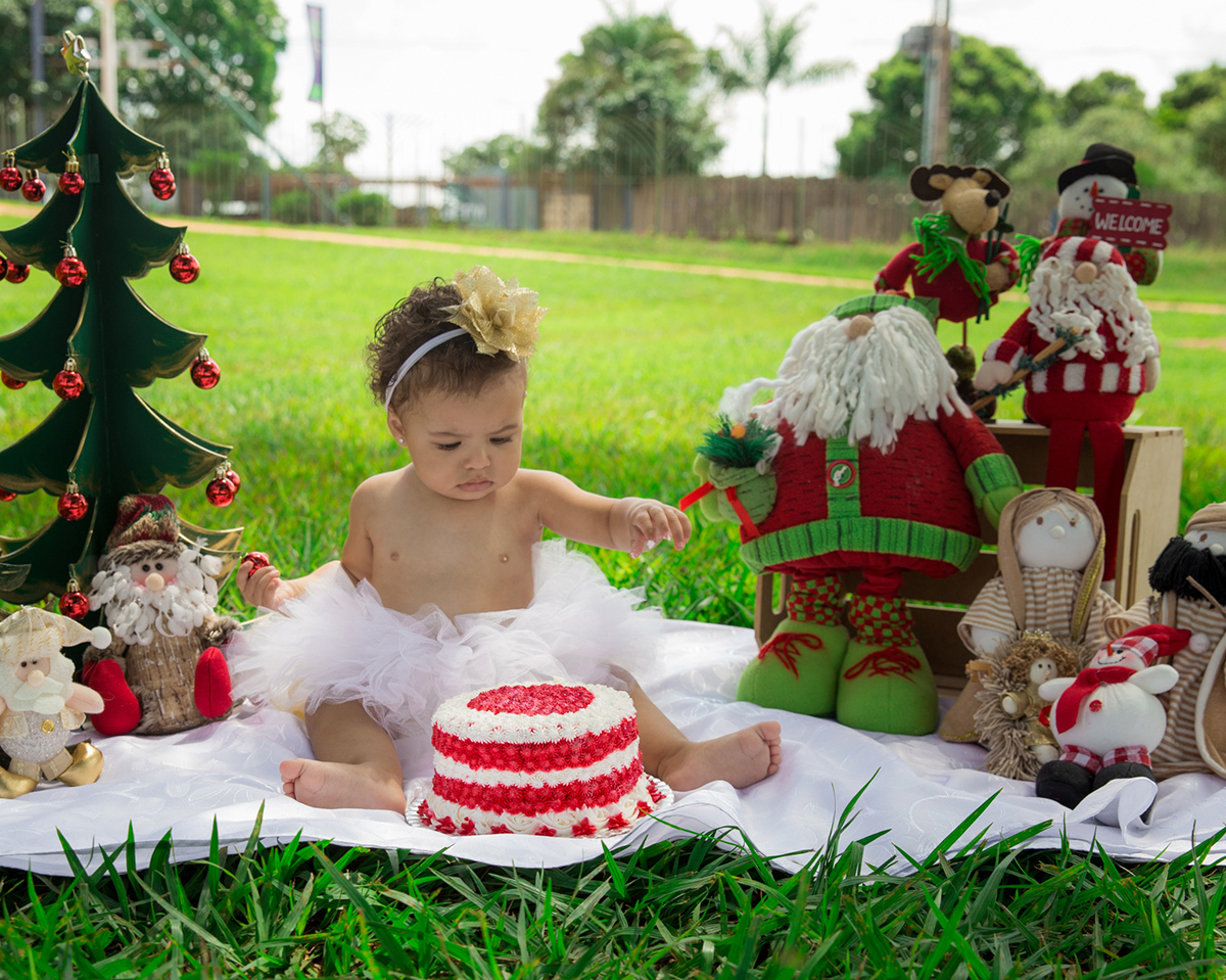 Bebê interage com cenário de natal, montado no gramado do parque, usa tutu branco e brinca com bolo vermelho