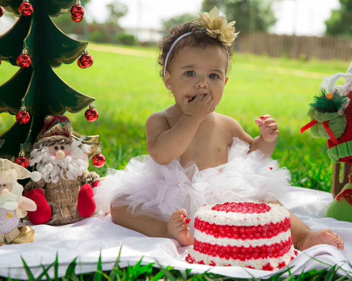 Bebê interage com cenário de natal, montado no gramado do parque, usa tutu branco e brinca com bolo vermelho, comendo glacê com as mãos