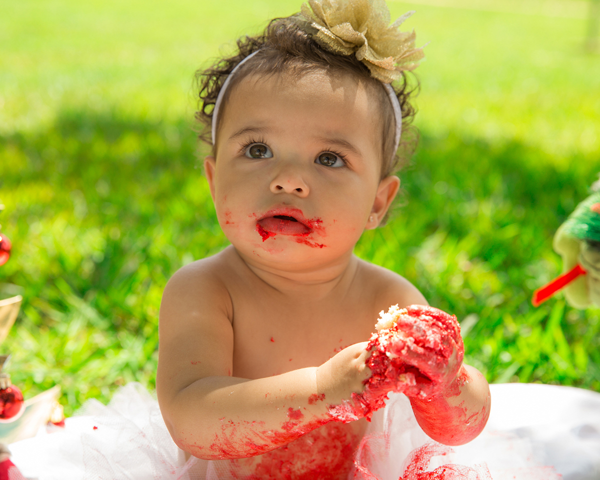 Bebê interage com cenário de natal, montado no gramado do parque, usa tutu branco e brinca com bolo vermelho, comendo glacê com as mãos