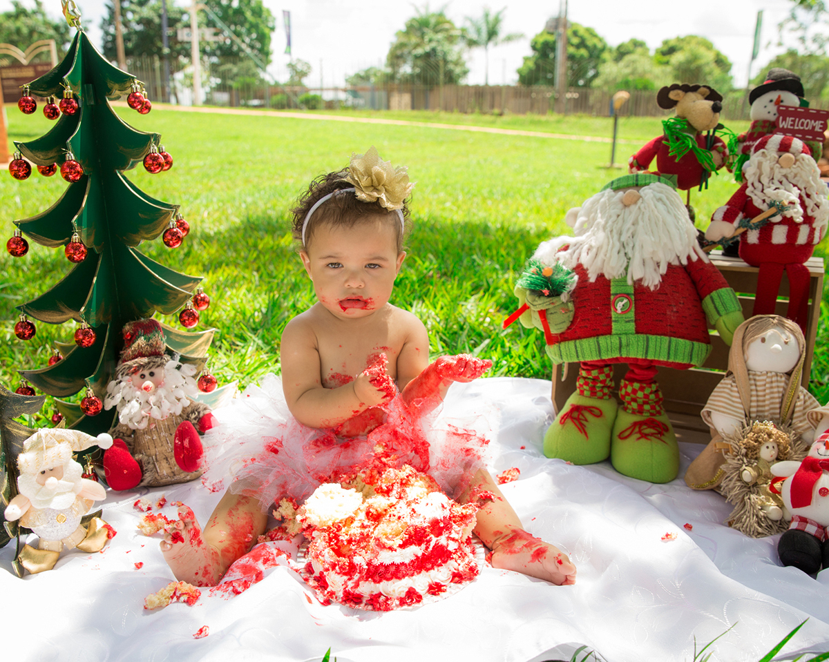 Bebê interage com cenário de natal, montado no gramado do parque, usa tutu branco e brinca com bolo vermelho, comendo glacê com as mãos. visão geral do cenário