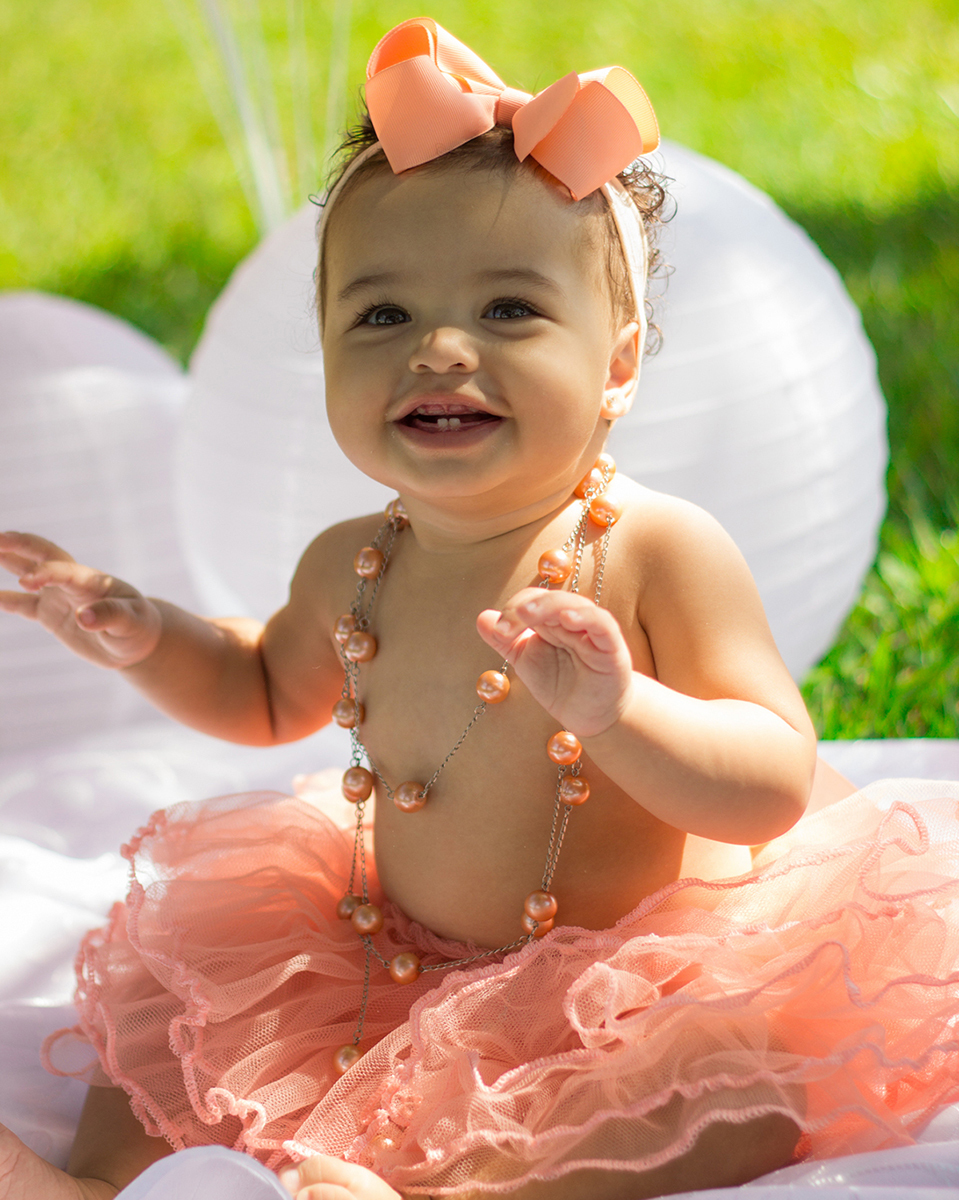 Bebê sentada no cenário, com saia de tutu, brinca e bate palmas