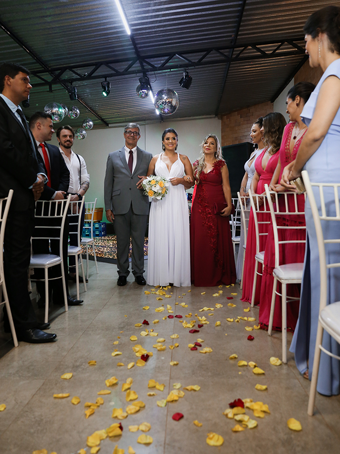 Entrada da noiva com os pais para cerimônia. Noiva de braços dados com o pai e a mãe. Foto feita no início do corredor, de baixo para cima