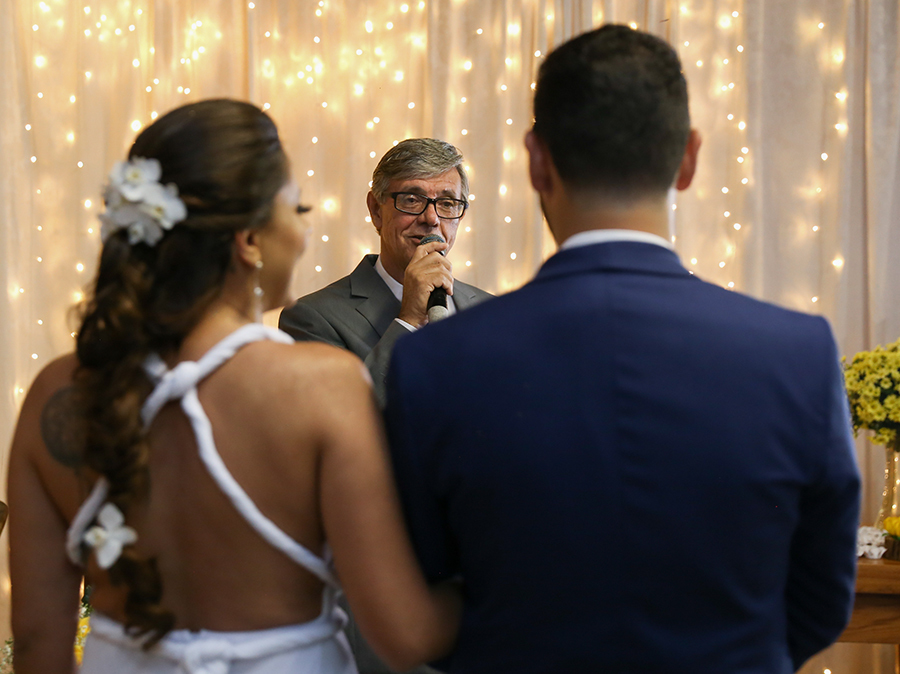 Detalhe do celebrante, pai da noiva. Visão das costas dos noivos, de braços dados, e detalhe do celebrante entre eles, mais ao fundo