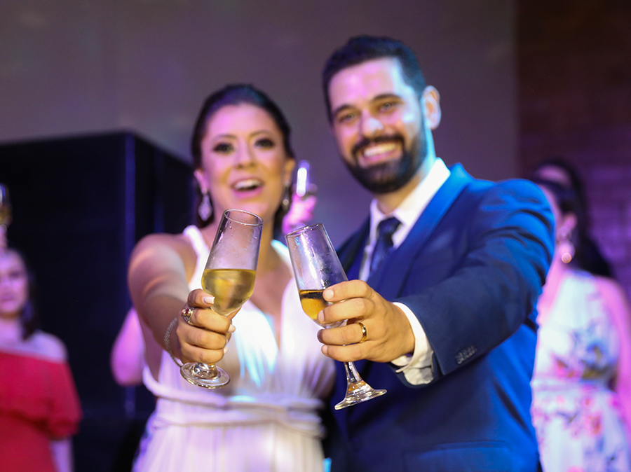 Noivos brindando. Copos no primeiro plano, ao fundo os noivos sorrindo