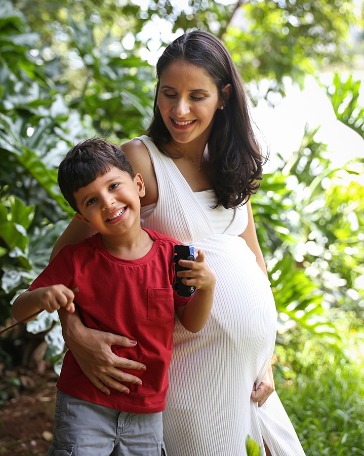 Gestante abraçada ao filho mais velho, olhando para ele. Filho olha para câmera, mostrando os carrinhos de brinquedo