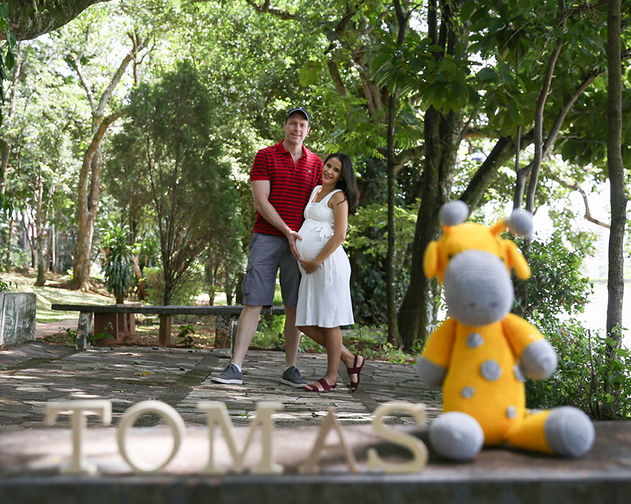 Papai e mamãe posam no mostrando a barriga em segundo plano. No primeiro plano, desfocados, girafa de pelúcia do bebê e letras formando nome Tomás