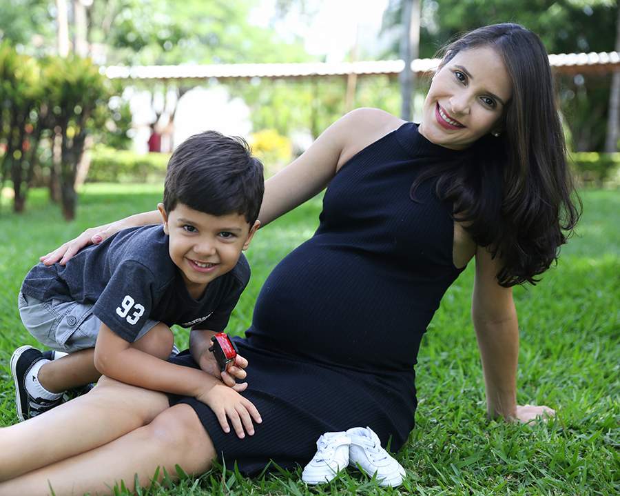 Mamãe sentada na grama, dando enfase a barriga, com sapatinhos do bebê ao lado, e irmão mais velho no colo da mãe