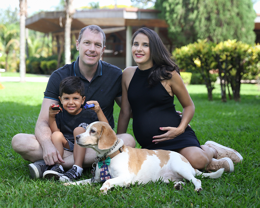 Toda a família, papai, mamãe, irmão e cachorro estão sentados na grama olhando para câmera.  Gestante com a mão da barriga