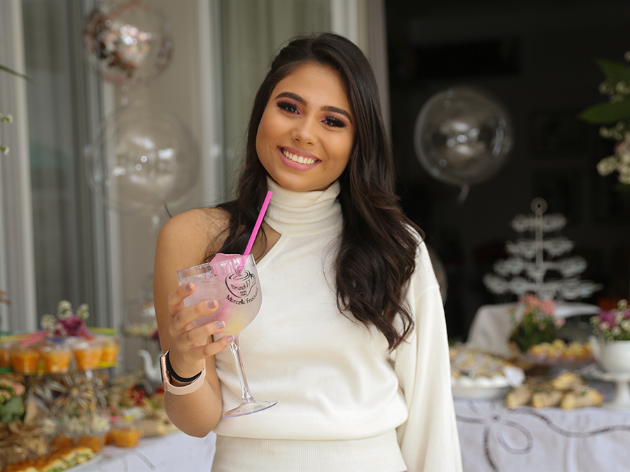 Aniversariante posa com a mesa do brunch ao fundo. Segura uma taça de drinks personalizada com seu nome