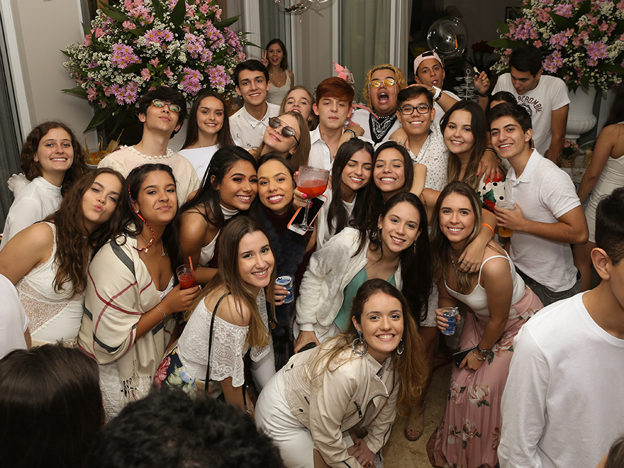 Aniversariante posa com seus amigos para foto, após o parabéns. Vista de cima em que aparecem uns 20 convidados posando para foto