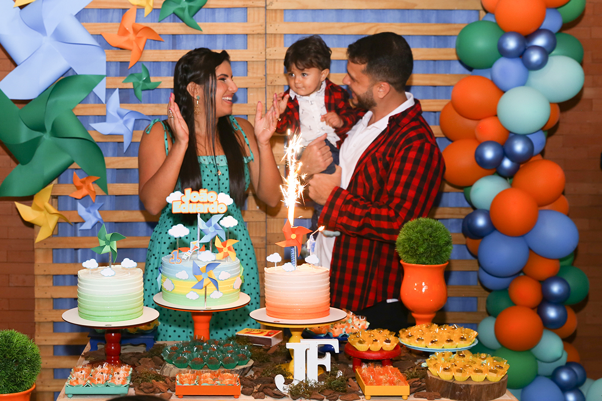 Paipai e Mamãe, com aniversariante no colo, atrás da mesa do bolo cantam parabéns junto com os convidados