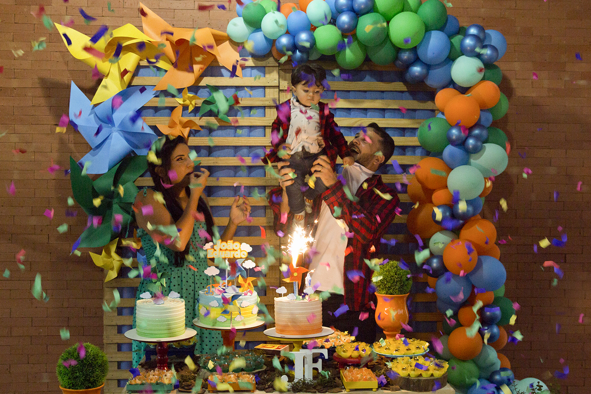 Paipai e Mamãe, atrás da mesa do bolo cantam parabéns junto com os convidados. Papai levanta aniversariante no alto enquanto chuva de papel cai