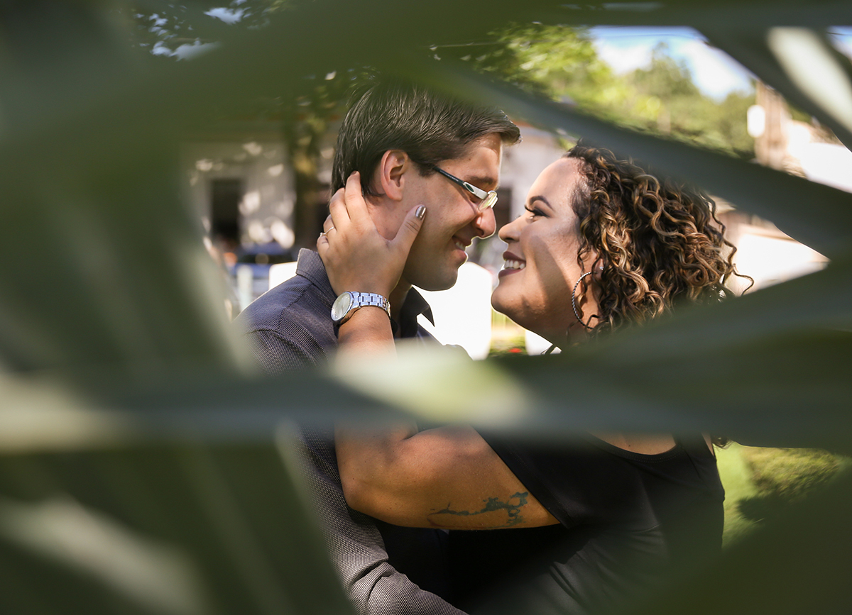 Casal, se abraça e se olha, em segundo plano. Em primeiro plano folhagem faz moldura desfocada
