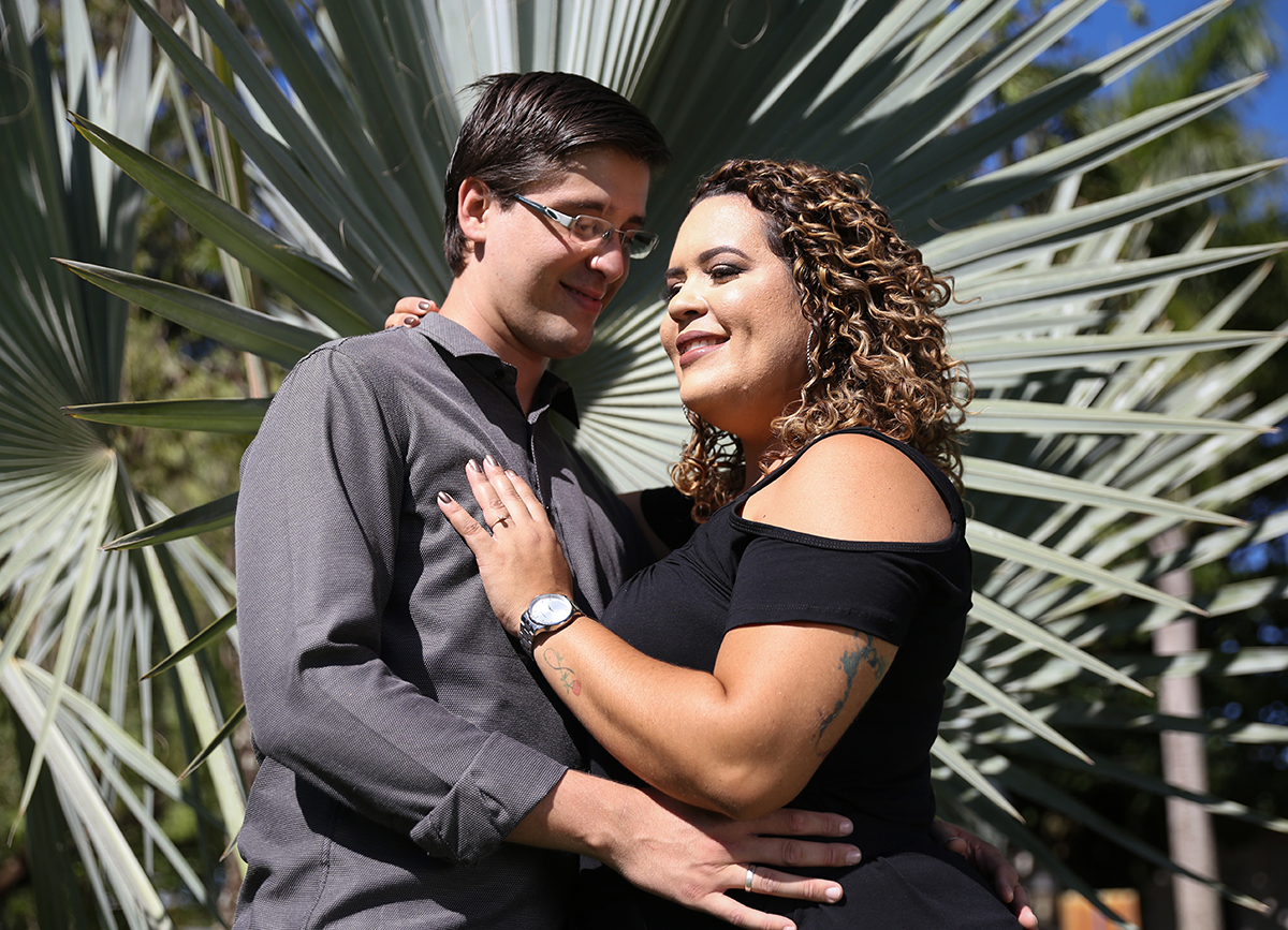 Casal se abraça, no primeiro plano, com folhagem fazendo moldura atrás