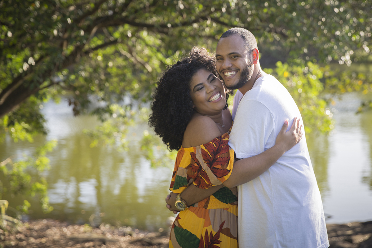 Casal abraçado, com rosto colado sorrindo. Ao fundo lago