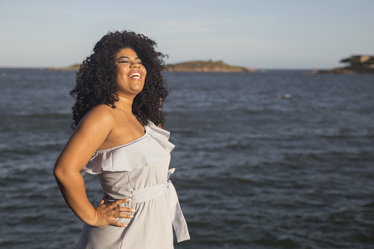 modelo sorrindo, com sol no rosto. ao fundo o mar