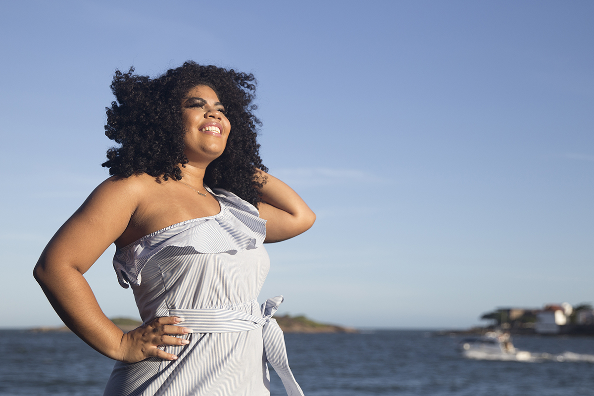 modelo sorrindo, com sol no rosto e a mão no cabelo. ao fundo o mar
