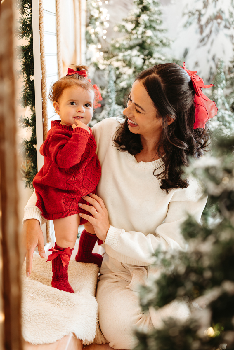 mãe vestido roupa clara, com cabelos pretos e detalhe de laço vermelho atras do cabelo. a mãe esta olhando sua filha que veste vestido vermelho
