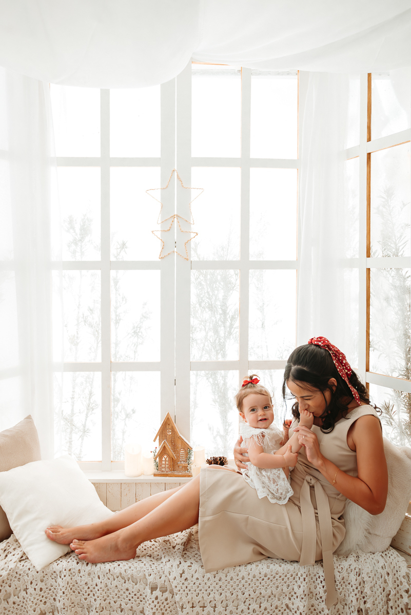 mãe e filha em cenário de natal clean. mãe esta sentada e esta dando um beijinho no pé de sua filha