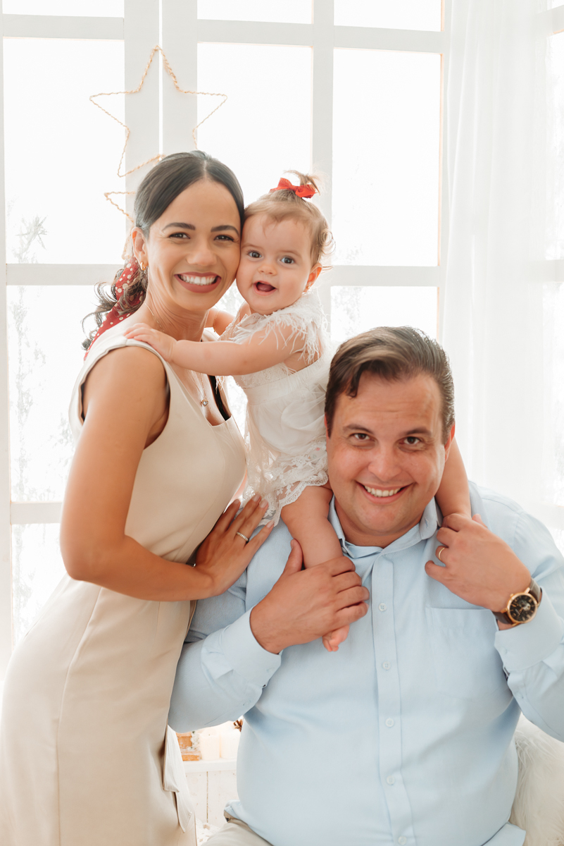 família em cenário de natal branquinho. filha esta no ombro do pai e mãe esta ao lado abraçando a bebe