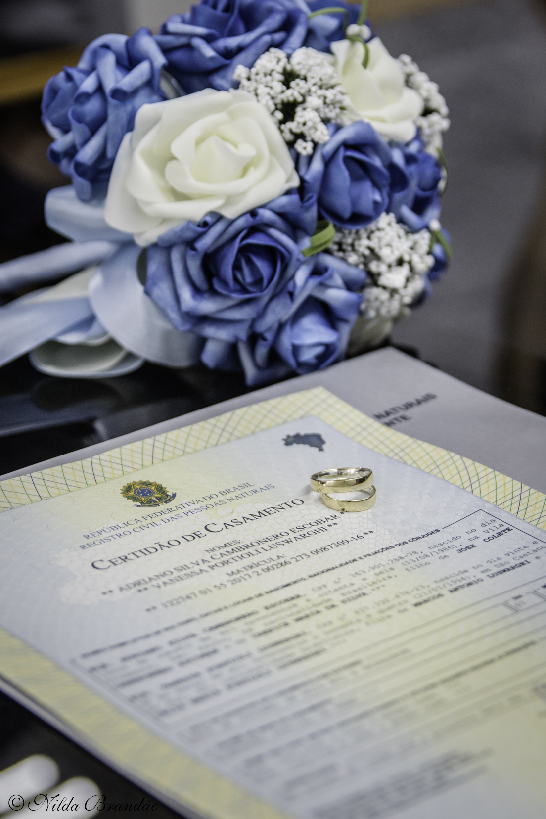 As alianças, fotografada encima da certidão de casamento, com o buquê como fundo. 