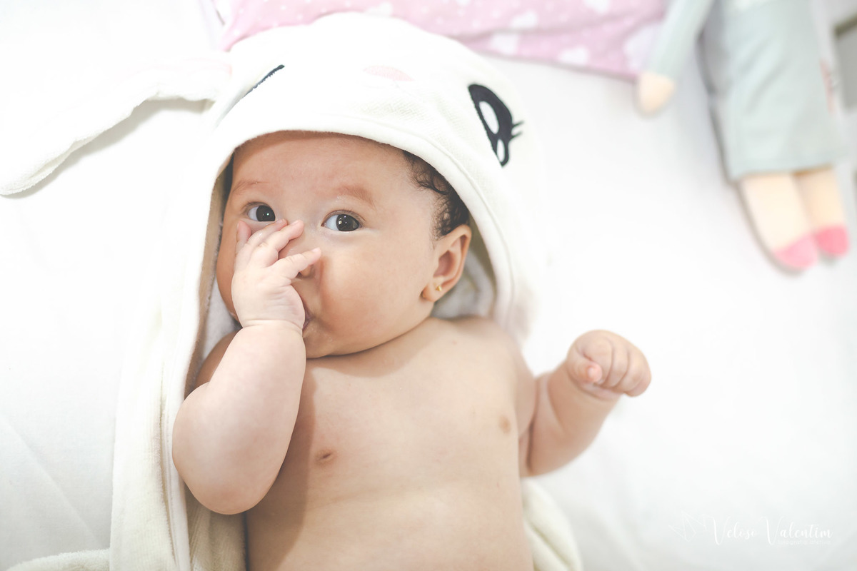 ensaio acompanhamento do primeiro ano do bebê Laura 3 meses em casa com a família ensaio lifestyle cotidiano Brasília DF Veloso Valentim fotografia afetiva bebê oriental com toalha de capuz com a mão na boca
