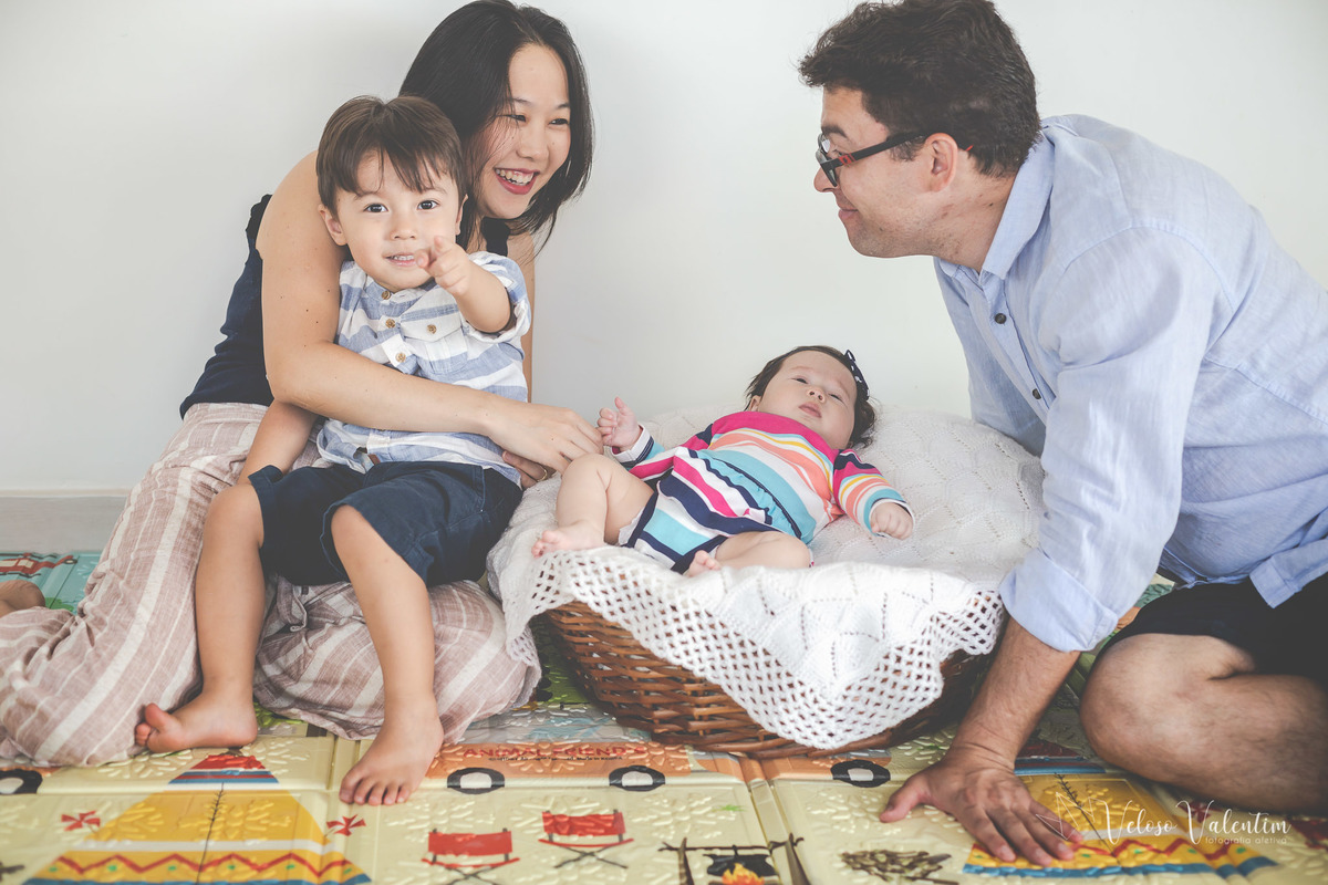 ensaio acompanhamento do primeiro ano do bebê Laura 3 meses em casa com a família ensaio lifestyle cotidiano Brasília DF Veloso Valentim fotografia afetiva família oriental pai e mãe se olham bebê deitada e criança sorrindo