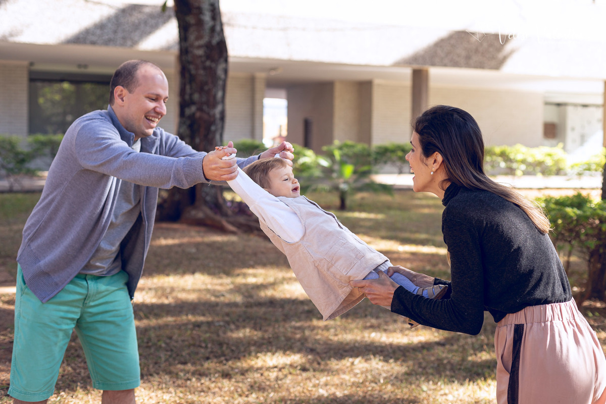 ensaio acompanhamento do primeiro ano do bebê Aurora 12 meses em casa com a família ao ar livre aprendendo a andar primeiros passos Brasília DF Veloso Valentim fotografia afetiva bebê sendo balançada pelos pais