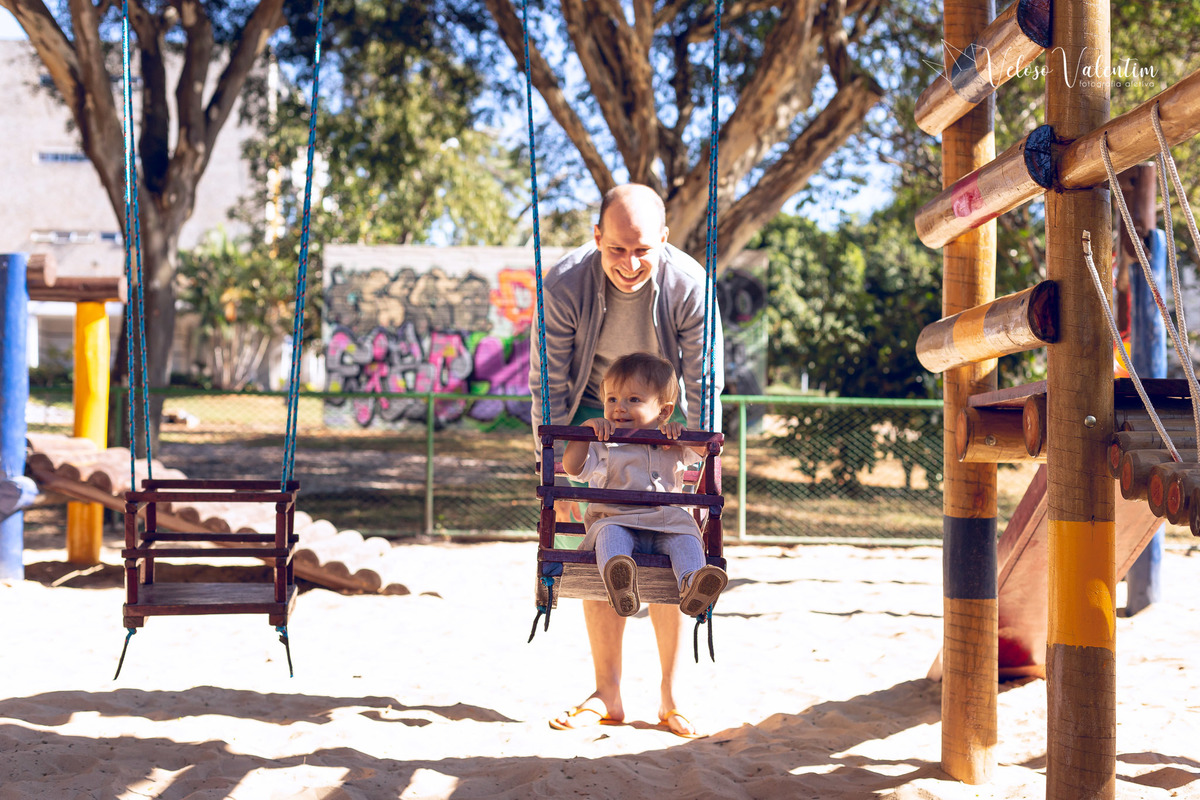 ensaio acompanhamento do primeiro ano do bebê Aurora 12 meses em casa com a família ao ar livre aprendendo a andar primeiros passos Brasília DF Veloso Valentim fotografia afetiva bebê no balanço do parquinho com o pai