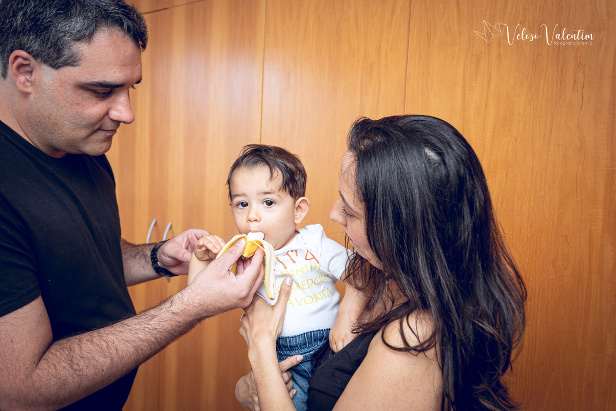 ensaio acompanhamento do primeiro ano do bebê Gabriel 12 meses em casa com a família ao ar livre engatinhando pai mãe avós ensaio família acompanhamento mensal fotográfico Brasília DF comendo banana