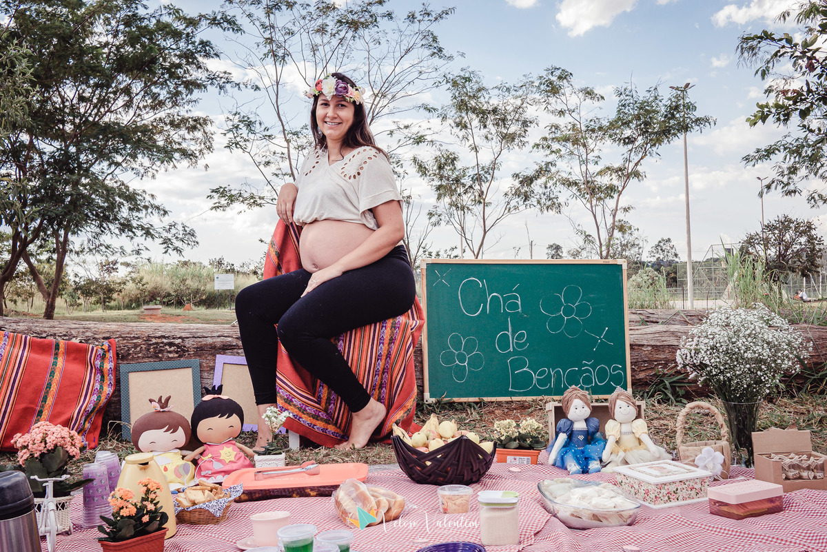 gestante posando sentada ao lado de uma placa escrito chá de bençãos com árvores ao fundo