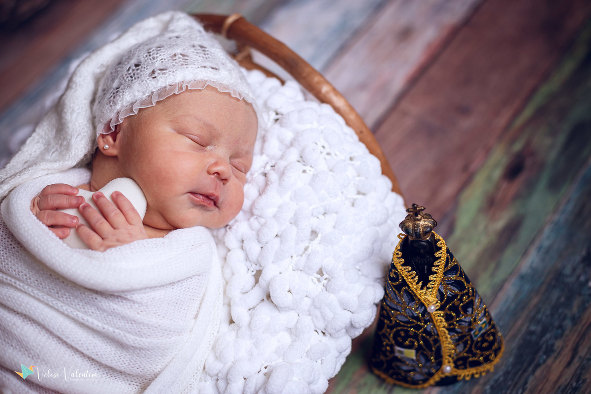 bebê deitada dormindo abraçada a um coraçãozinho ao lado de uma imagem de nossa senhora ensaio newborn em casa Brasília